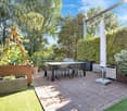 A modern outdoor patio with a dining table, four chairs, and a large umbrella. The area is surrounded by greenery, including potted plants and a wooden trellis, with a wooden deck and a view of trees in the background.
