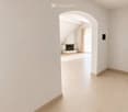 Empty hallway with beige tiled flooring and white walls, featuring an arched doorway leading to another room with a fireplace and window.