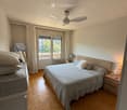 Cozy bedroom with wooden herringbone flooring, large double bed with pillows and blankets, nightstand with lamp, ceiling fan, and large window with curtains leading to a balcony.