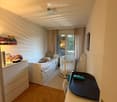 Cozy bedroom with a single bed, baby crib, changing table, and a shelf with books and folded clothes. The room features wooden flooring and large windows with curtains.