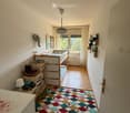 A cozy children's bedroom with a colorful rug, a bunk bed, a small table, and a storage unit. The room features a large window with curtains and a view of greenery outside.