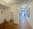 Bright hallway with wooden flooring, white walls, and modern lighting fixtures. Features a mirror, small decorative table, and artwork on the walls.