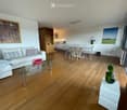 Modern living room with light wooden flooring, white sofa, glass coffee table, dining area with glass table and chairs, and decorative artwork on the walls.