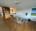 Modern dining area with a glass table and transparent chairs, a kitchen counter, and a wooden cupboard with shelves.