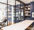 Modern office space with dark blue walls, white tables, chairs, shelves filled with books and decorative items, and a laptop on a desk.