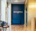 Modern hallway with wooden flooring, dark blue accent wall with 'enigma' text, two contemporary chairs, small round table with a book and vase, and a glass door leading to a view of trees.