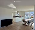 Open-plan living and dining area with a large wooden dining table, modern chairs, a kitchen area with white cabinets, and a large TV screen on the wall.