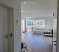 Modern living room with white walls, wooden floor, large windows, sofa, ottoman, fan, and shoe rack near the entrance.