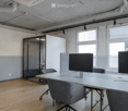 Modern office space with wooden flooring, large windows, white table, grey chairs, and two screens on the table.