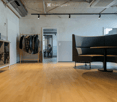 Modern, minimalist room with light wooden flooring, a large L-shaped sofa, a small round table, a wooden shelf, and a coat rack with jackets.