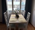 Dining area with a wooden table, four chairs, placemats, and a potted plant near a large window with blinds.