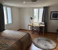 Cozy bedroom with a wooden bed, patterned bedspread, a round shaggy rug, a wooden desk with a chair, a desk lamp, and a printer on the desk. Large windows with curtains and a picture frame on the wall.