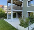 Modern residential building with a raised ground floor, balcony, and well-maintained garden area with grass, plants, and a paved patio.