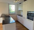 Modern kitchen with white cabinets, black countertops, a sink with a faucet, and an oven. The space features wooden flooring and a window with a view of greenery.