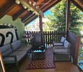 A cozy balcony with a wooden ceiling, a sofa, two chairs, a coffee table, and a small round table with cups and a teapot. The balcony has a patterned rug and overlooks a green, tree-filled area.