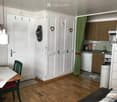 A compact, well-lit room with a wooden floor, white walls, and white cabinetry. A small dining area features a wooden table with a red and white tablecloth and a green chair. A kitchen area includes a sink, microwave, and a small refrigerator. The room ha