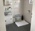 A small bathroom with beige tiled walls and floor, a white sink with a mirror above it, a toilet with a blue seat cover, a white cabinet under the sink, a gray bath mat, and a towel hanging on the wall.