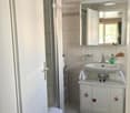 Modern bathroom with white tiles, a sink with a mirror above, a small cabinet, and a shower stall. A towel is placed on the floor.