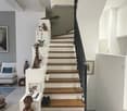 Modern hallway with a staircase featuring wooden steps and white risers, black metal railing, decorative items including sculptures, framed pictures, and a potted plant on a white console table. The hallway has white walls, a sleek design, and a view into