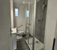Modern bathroom with a glass-enclosed shower, toilet, and sink. The space features tiled walls and a small window for natural light.