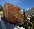 Modern wooden house with a flat roof, situated in a scenic mountainous area with a view of a snow-capped peak and a town below.