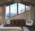 Modern bedroom with large windows offering a mountain view, featuring a bed, a chair, a table, and a lamp.