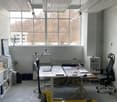 Office space with two desks, chairs, laptops, a fan, a refrigerator, and a screen on the wall.