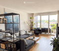 Modern living room with light wooden flooring, grey sofa, coffee table, bookshelf, and large windows offering a view of the city.