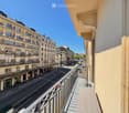 Balcony overlooking a quiet urban street lined with classic European buildings, featuring metal railings and wooden flooring.