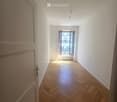 Empty hallway with wooden herringbone flooring, white walls, and a single open door leading to a balcony area with a radiator visible.