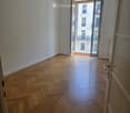 Empty room with wooden herringbone flooring, white walls, and an open door leading to a balcony with a view of a building.