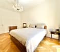 Elegant bedroom with a wooden bed, nightstand, mirror, and chandelier. The room features light-colored bedding and wooden flooring.