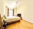 Cozy bedroom with wooden herringbone flooring, a large bed with white bedding, a nightstand with a lamp, a mirror, and a television on a wooden stand. A guitar is placed near the window, and the room features a chandelier and large windows with curtains.