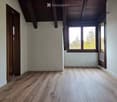 Empty room with wooden flooring, white walls, and large windows letting in natural light. The room features exposed wooden beams on the ceiling and a partially open door leading to another space.