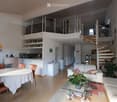 Open-plan living and dining area with white walls, wooden flooring, a white sofa, glass coffee table, dining table with chairs, kitchen area, and a spiral staircase leading to a lofted area.