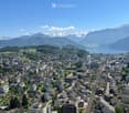 Aerial view of a town nestled in a valley with surrounding mountains and a lake in the background. The town features a mix of residential and commercial buildings, roads, and green spaces.