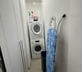 A small, well-lit room with a washing machine, an ironing board, and cleaning supplies on a shelf.