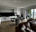 Modern, open-plan living and dining area with a leather sofa, round dining table, chairs, and a kitchen with white cabinets and a dishwasher. Large windows offer a scenic view.