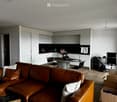 Modern living and dining area with a brown leather sofa, a laptop on the sofa, a round dining table with chairs, a kitchen area with white cabinets and a black countertop, and an office area with a desk and chair.
