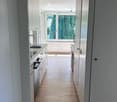 Bright, modern hallway with light wooden flooring, white walls, and built-in cabinets on both sides. A large window at the end offers a view of greenery outside.