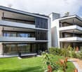 Modern two-story house with white and grey exterior, large windows, balconies, and a well-maintained lawn.