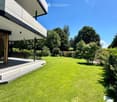 Well-maintained lawn with trimmed hedges and trees, modern house with large windows and raised ground floor visible in the background.