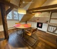 A cozy attic kitchen with wooden beams, a round wooden dining table with four chairs, white cabinets, a black oven, and a hanging pendant light.