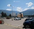 Parking lot with several cars, surrounded by trees and mountains, with commercial buildings and flags visible in the background.