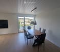 Modern dining room with wooden floor, large window offering a scenic view, a dining table with chairs, and a flat-screen TV mounted on the wall.