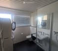 Modern bathroom with a glass-enclosed shower, sink, toilet, and mirrored cabinet. Features include a window with blinds, a towel rack, and a washing machine visible through the mirror.