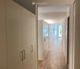 Modern hallway with wooden flooring, white walls, and built-in storage units with sleek handles. Natural light streams in from the end of the corridor.