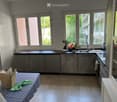 Modern kitchen with grey cabinets, tiled floor, and large windows. Features a sink, faucet, oven, and a small table with a green cloth.