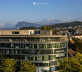Modern multi-story building with large glass windows and a curved facade, surrounded by trees and with a scenic mountain backdrop.