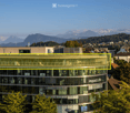 Modern multi-story building with large glass windows and a curved facade, featuring a rooftop terrace with outdoor seating and a scenic mountain backdrop.